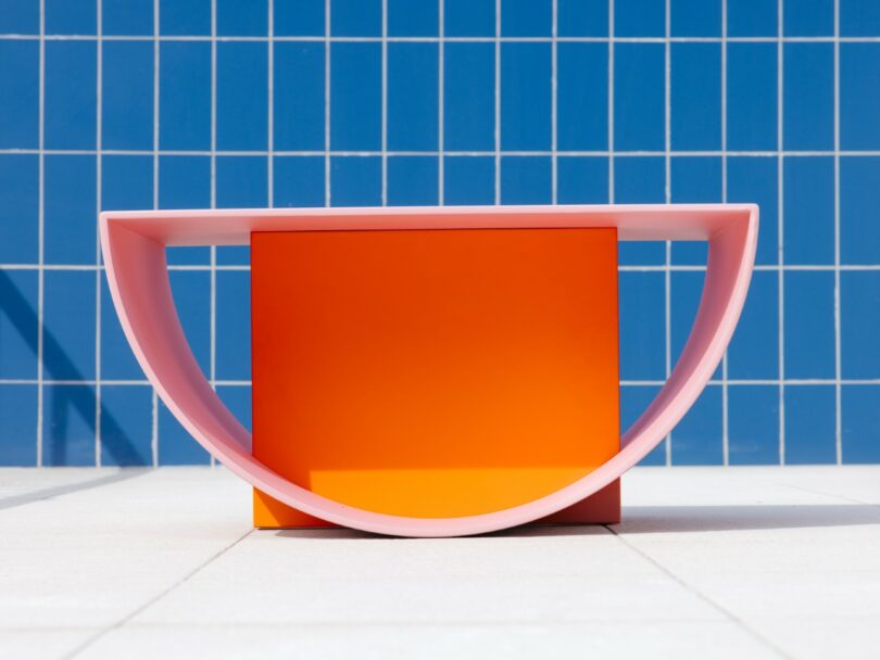 A geometric table with a pink semicircular frame and orange rectangular base stands on a white tiled floor against a blue tiled wall