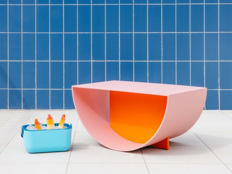 A blue cooler with popsicles and a modern pink-orange semicircular table sit on white tiles against a blue tiled wall