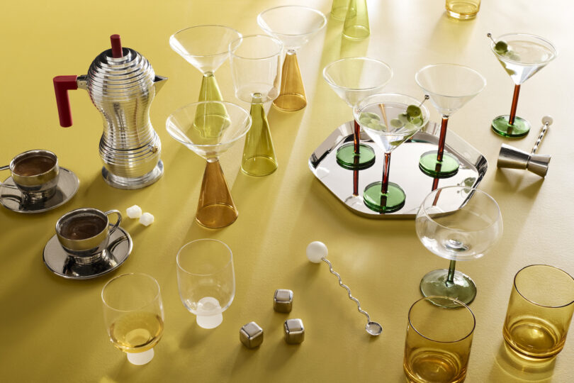 A variety of glassware, cups, a metal coffee maker, and cocktail accessories are arranged on a yellow surface with a silver tray and several drinking glasses containing drinks and olives