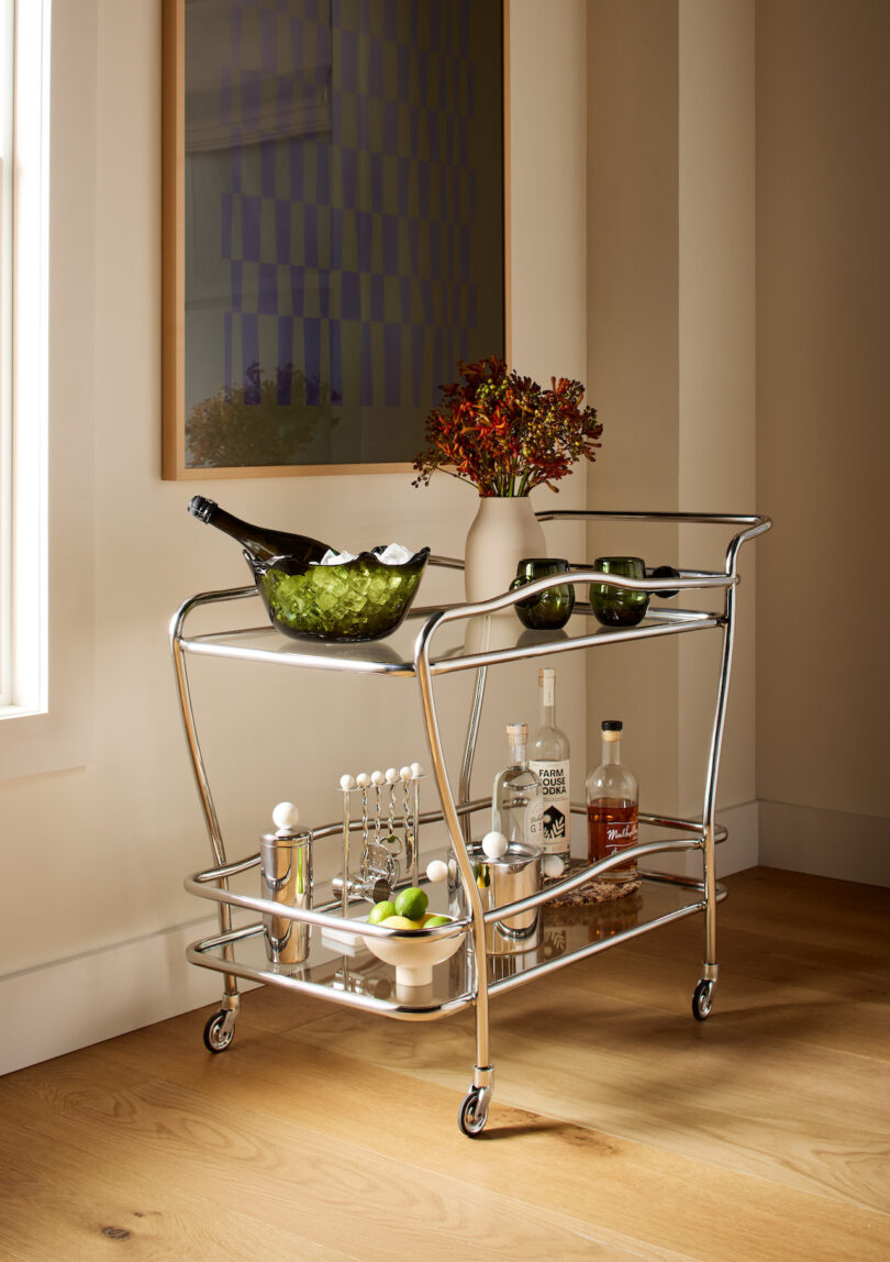 A chrome bar cart with bottles, cocktail tools, glasses, and a bowl of ice with champagne sits by a window and wall art on a wooden floor