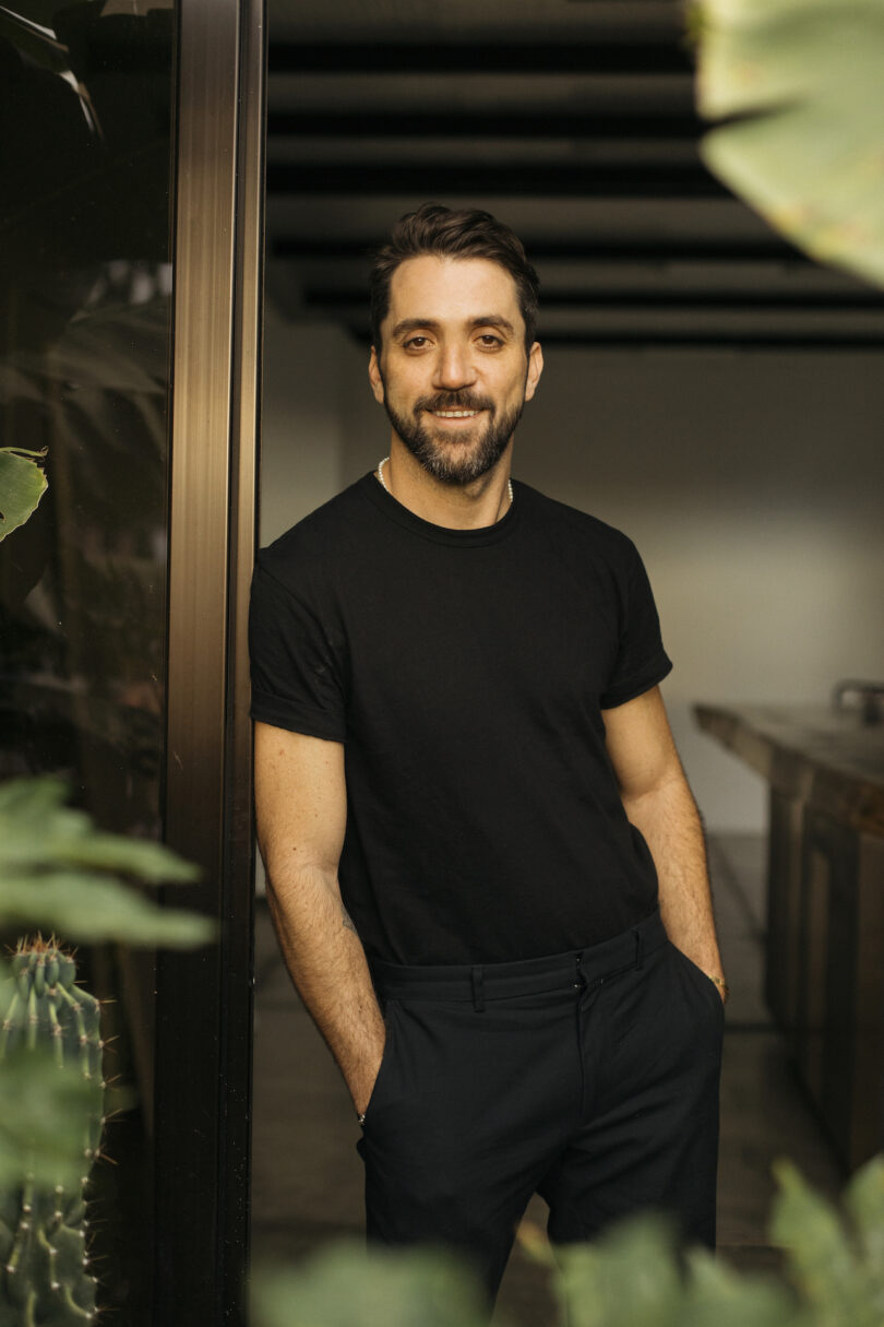 A man with a beard and mustache stands indoors, leaning against a door frame, wearing a black t-shirt and pants, with hands in his pockets, surrounded by green plants