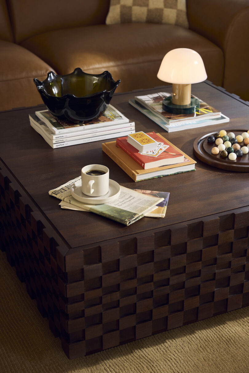 A dark wood coffee table with a cup of coffee, books, a lamp, a decorative glass bowl, magazines, and a tray of colorful spheres, set in a living room with a brown couch