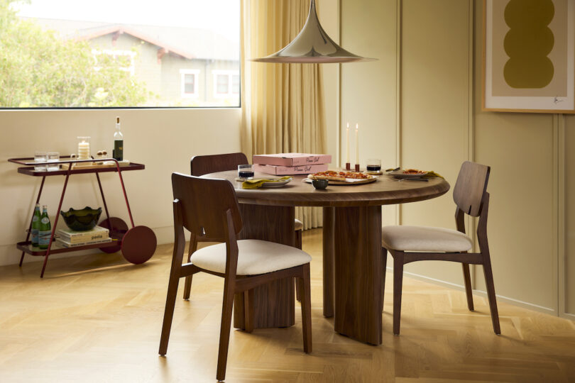 A modern dining room with a round wooden table set for two, a pink pastry box, and a bar cart near a window with light curtains