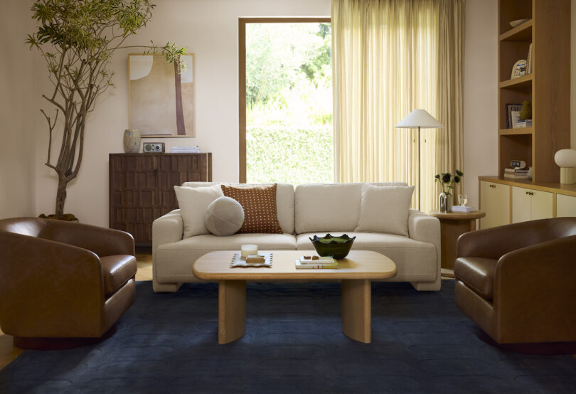 A modern living room with a white sofa, two brown armchairs, a wooden coffee table, blue rug, large window, and decorative items on shelves and tables