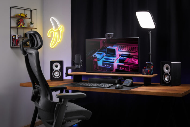 A modern gaming and streaming setup featuring a large monitor, speakers, microphone, webcam, and Elgato LED panel light on a wooden desk, with a neon banana light and shelves in the background.