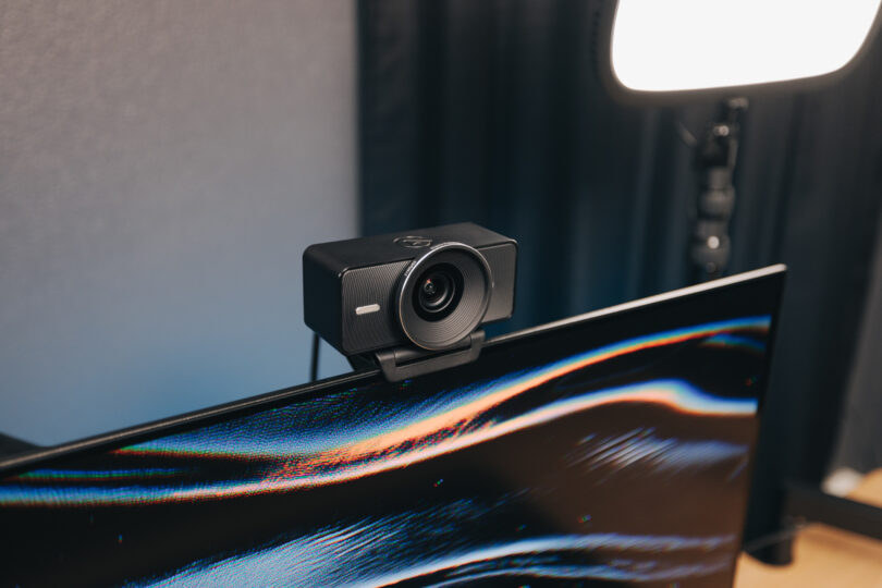 An Elgato black webcam is mounted on top of a computer monitor, with a bright studio light visible in the background.