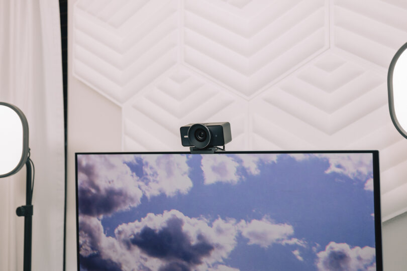 An Elgato webcam is mounted on top of a monitor displaying a cloudy sky, with two softbox lights on either side and geometric acoustic panels in the background.