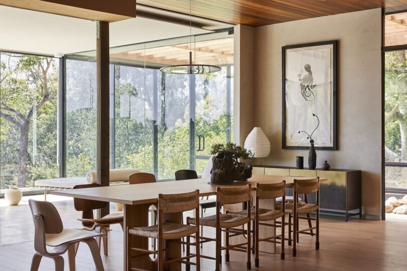A modern dining room with a wooden table, six chairs, decorative plants, large glass windows, and a framed artwork on the wall.