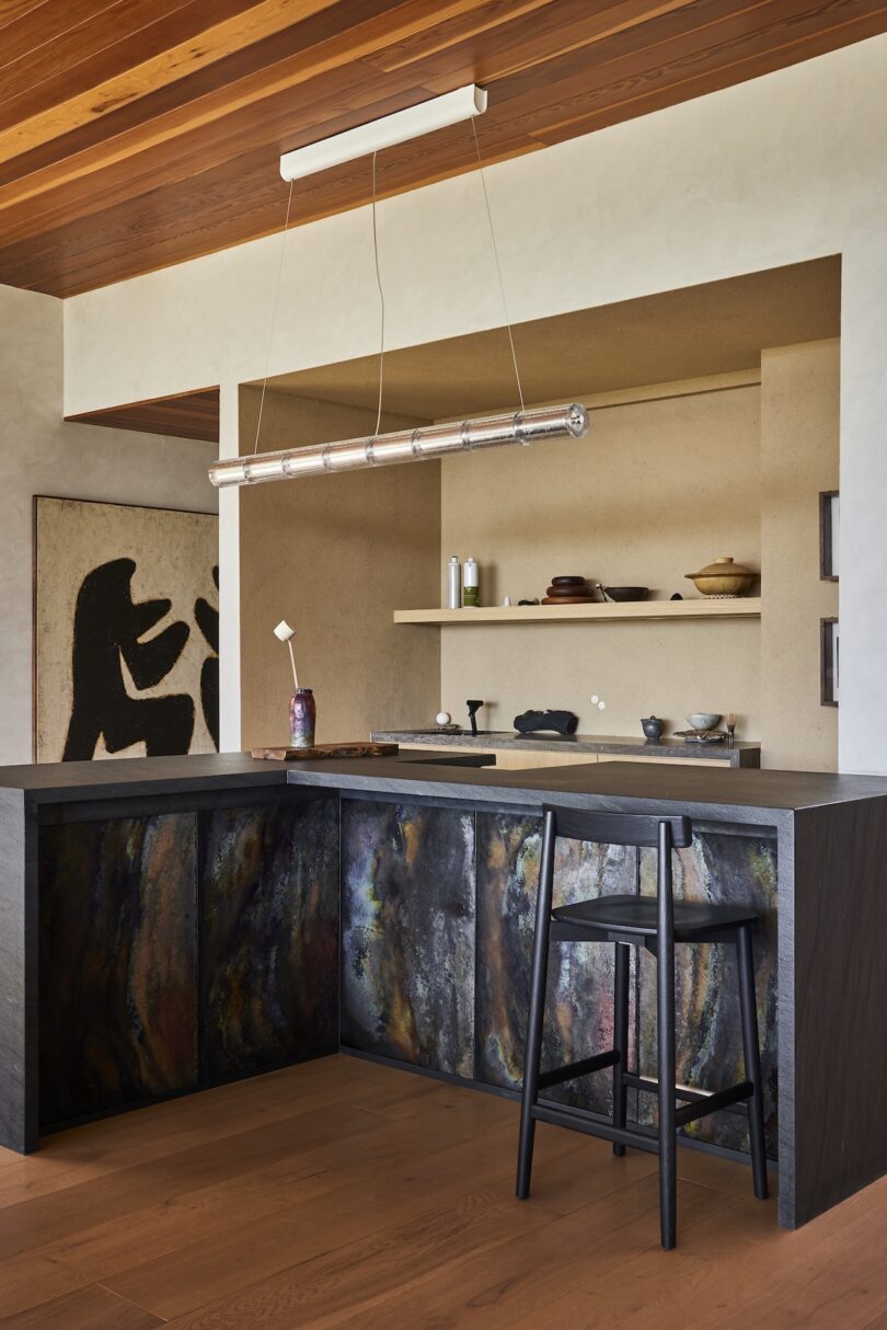 Modern kitchen with dark, textured cabinetry, a black barstool, open shelves holding ceramics, wood ceiling, and abstract wall art in the background.