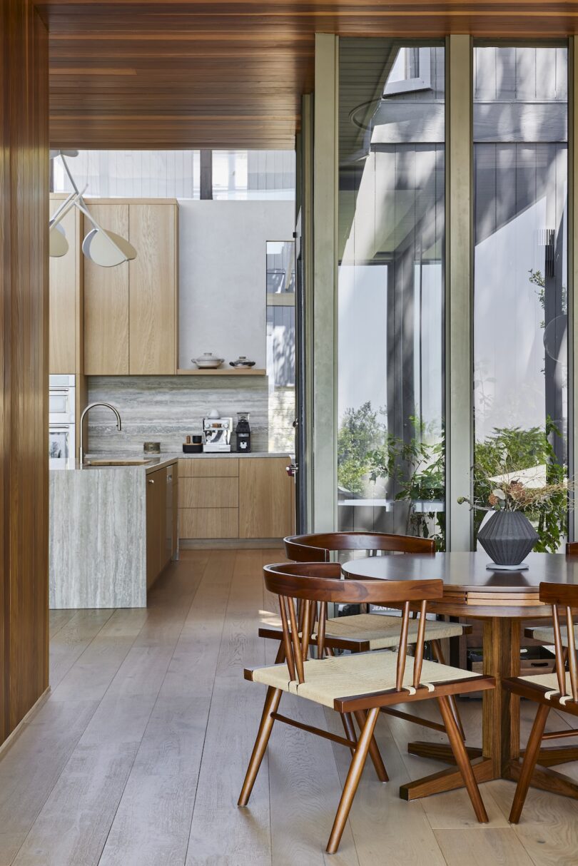 Modern kitchen and dining area with wooden furniture, large floor-to-ceiling windows, and natural light. Kitchen features light wood cabinets and marble countertops.