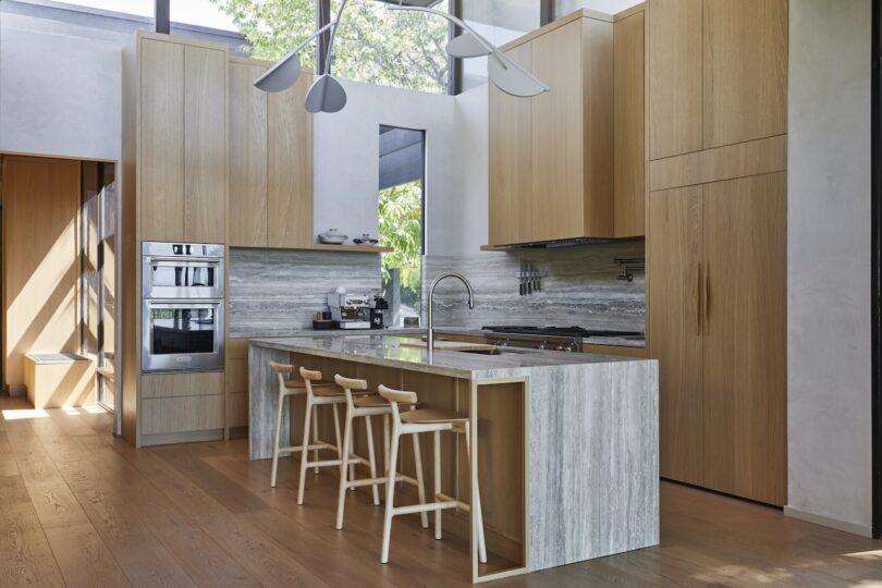 Modern kitchen with light wood cabinets, a marble island with three wooden stools, built-in appliances, and large windows letting in natural light.