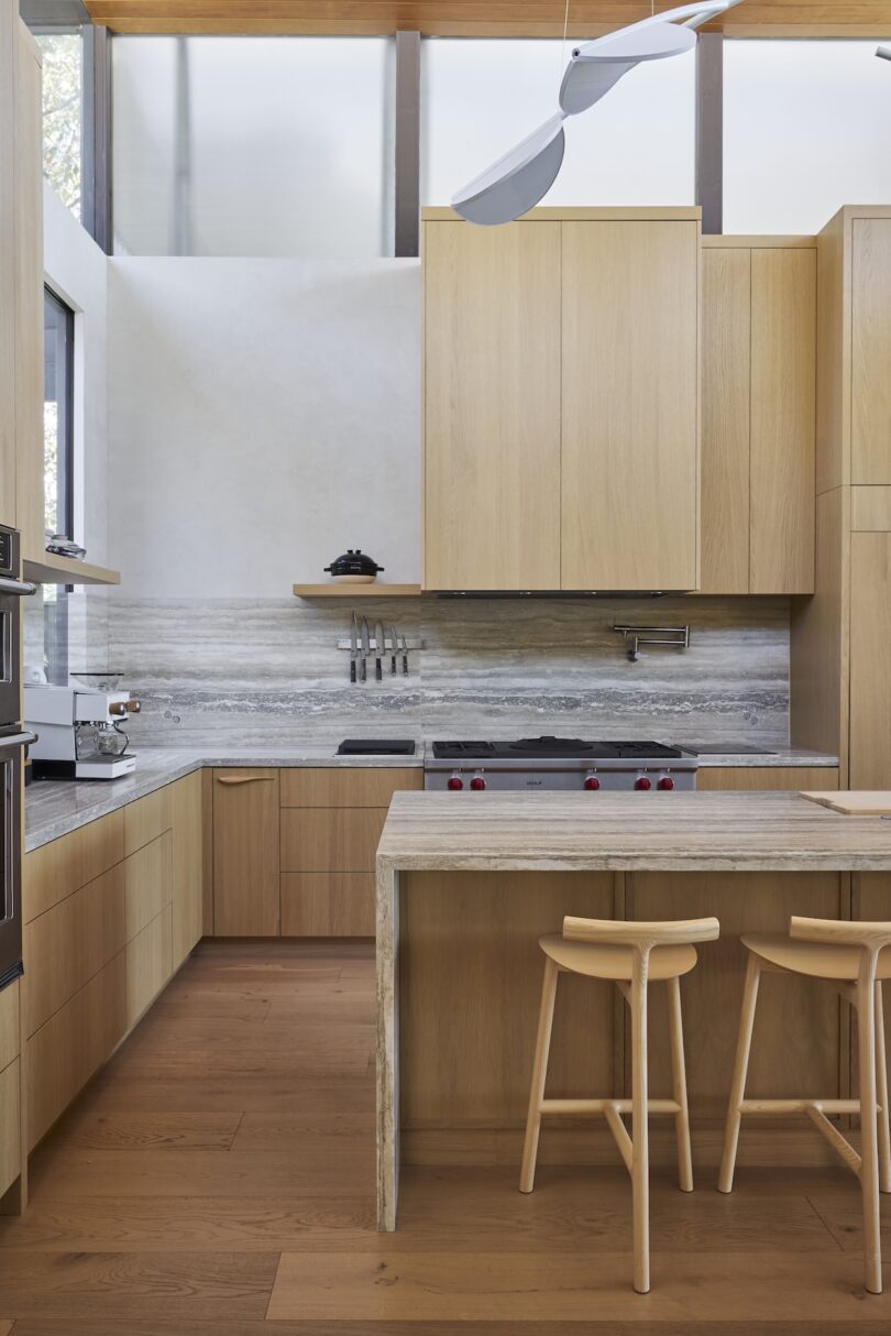 Modern kitchen with light wood cabinets, a marble backsplash, built-in appliances, and an island with two wooden stools under natural light.