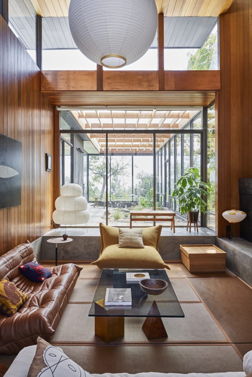 A modern living room with wood-paneled walls, a brown leather sofa, glass coffee table, large paper lantern light, and floor-to-ceiling windows overlooking a patio with plants.