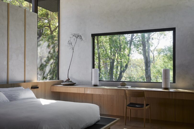 Minimalist bedroom with a large bed, wooden desk, single chair, tall vases, and large windows offering views of trees and greenery outside.