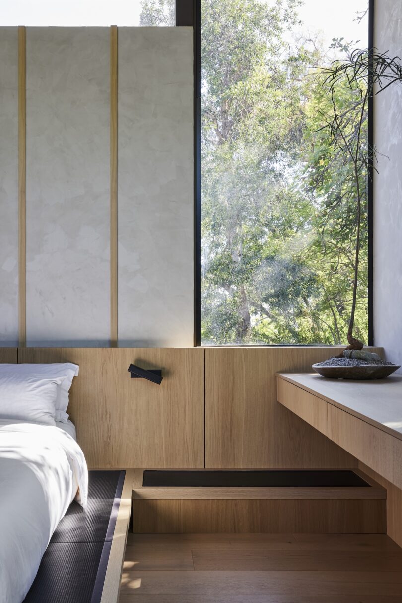 Minimalist bedroom with light wood paneling, a large window showing trees outside, a white bed, and a simple desk with a small plant and decorative items.