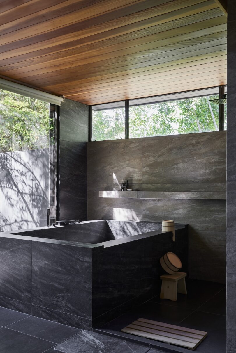 Modern bathroom with dark stone walls and a soaking tub, wooden ceiling, large window with greenery outside, and minimal decor including a stool and bath accessories.