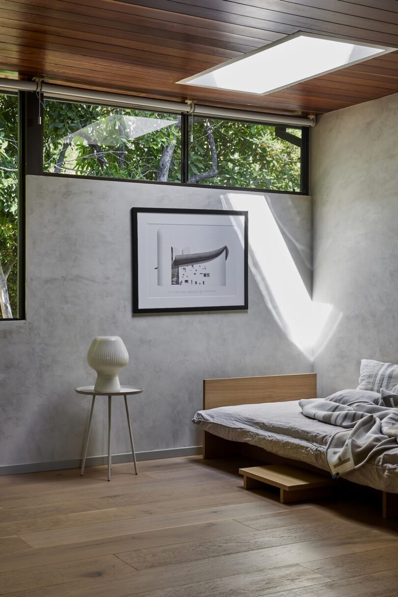 Minimalist bedroom with a low wooden bed, gray bedding, a small side table with a white vase, wall art, and large windows letting in natural light.