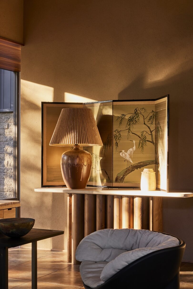 A large ceramic lamp with a pleated shade sits on a table next to a decorative folding screen with a bird motif; warm sunlight casts shadows on the wall.
