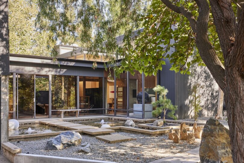 Modern house with large windows and a minimalist courtyard featuring gravel, rocks, small plants, and a tree in the foreground.