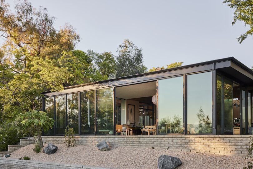 A modern house with large floor-to-ceiling glass windows sits on a gravel and stone landscape surrounded by trees and greenery.