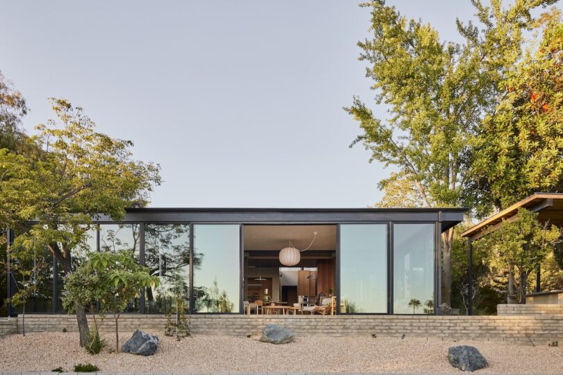 Modern single-story house with large floor-to-ceiling glass windows, surrounded by trees and gravel landscaping, revealing a view of the interior dining area.