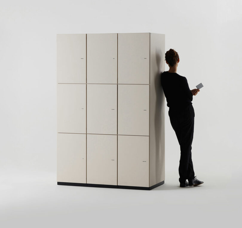 A person in black clothing leans against a beige locker unit while holding a rectangular object, against a plain white background.