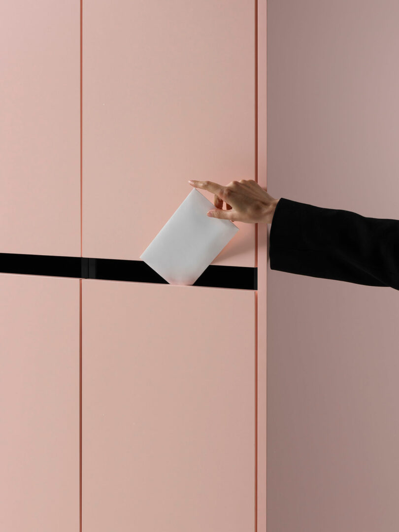 A person inserts a white envelope into a horizontal slot on a pink wall or panel, wearing a black sleeve.
