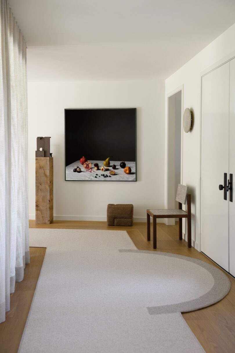 Minimalist hallway with light walls and flooring, featuring a wooden pedestal, abstract art, a chair, and a modern black-and-white fruit still life painting on the wall.