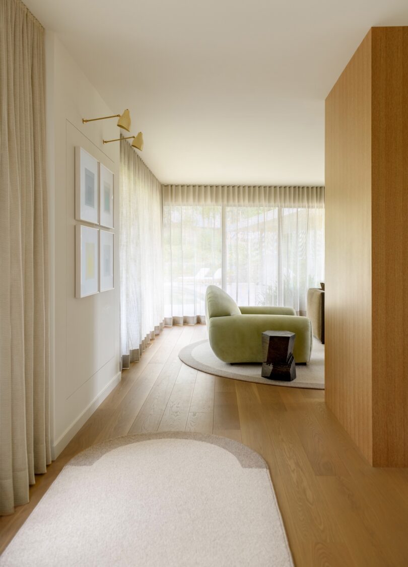 A modern living room with light wood floors, sheer curtains, green lounge chair, nested tables, and framed art on the wall. The space has minimal, soft-toned decor and natural light.
