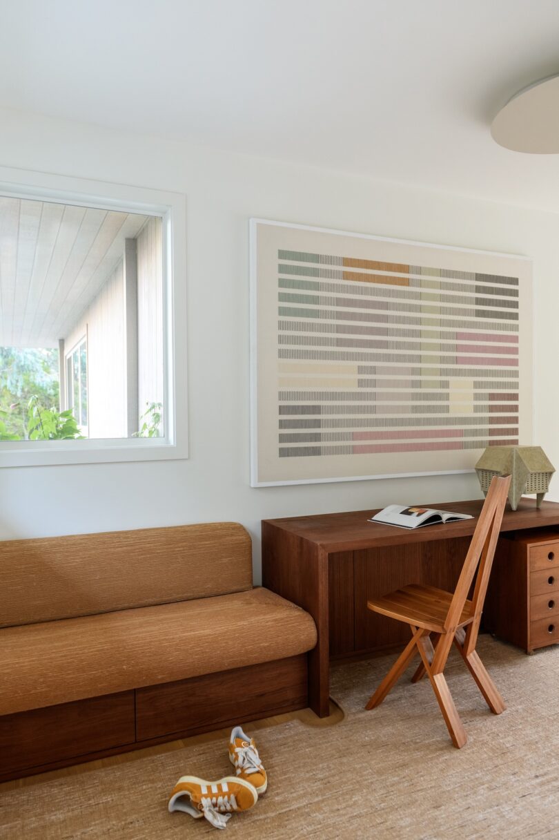 A minimalist study room with a wooden desk, chair, sofa, abstract wall art, open book, and a pair of yellow sneakers on a beige carpet.