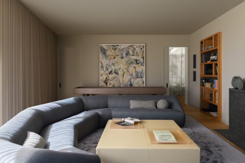 Modern living room with a curved gray sofa, a beige square coffee table, abstract wall art, built-in bookshelves, and neutral-colored decor.