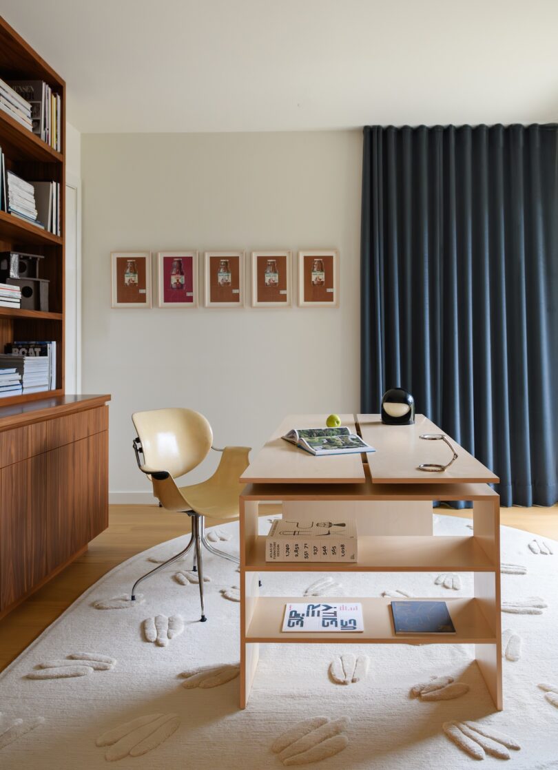 A modern home office with a wooden desk, a beige chair, bookshelves, framed art on the wall, and dark curtains covering a window.