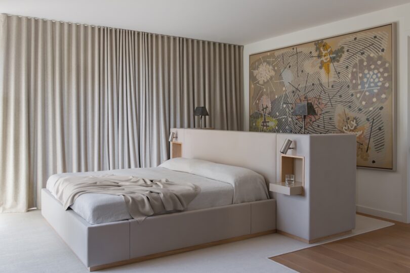 A modern bedroom with a low platform bed, built-in shelves, neutral bedding, floor-to-ceiling curtains, and a large abstract artwork on the wall.