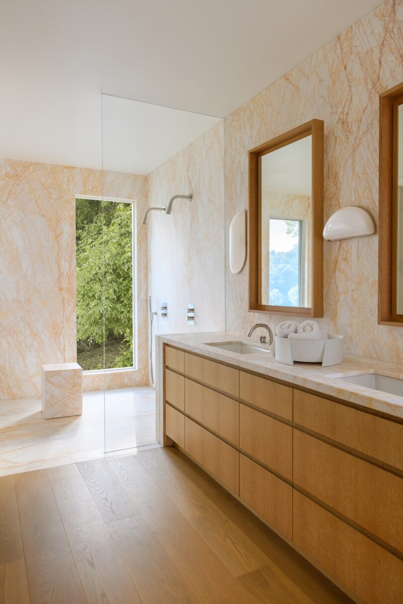 A modern bathroom with light wood cabinets, marble walls, dual sinks, a glass-enclosed shower, and a large window showing greenery outside.