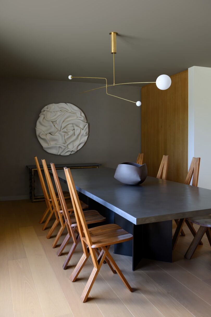 A modern dining room features a black rectangular table with six wooden chairs, a minimalist light fixture, and abstract wall art. A decorative bowl sits at the table's center.
