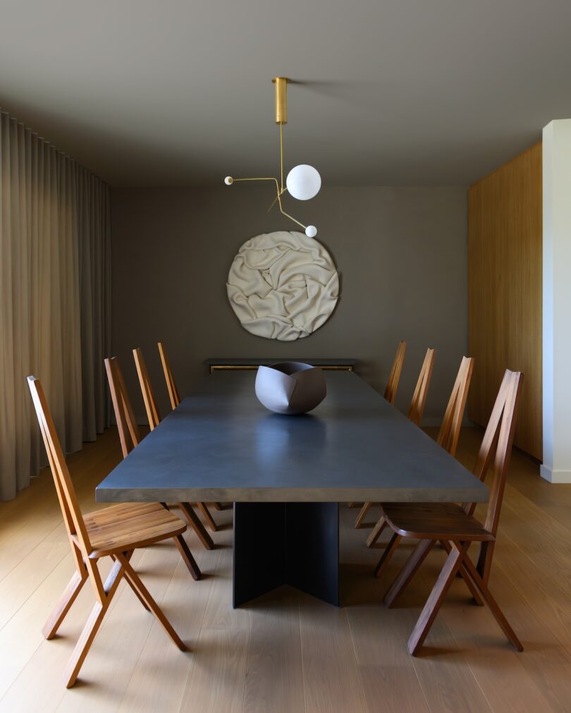 A modern dining room with a dark rectangular table, eight wooden chairs, a sculptural bowl centerpiece, abstract wall art, and a minimalist pendant light.