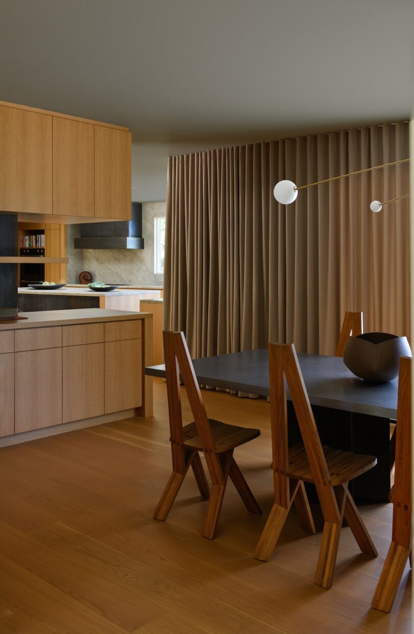 A modern dining area with a black table, four wooden chairs, wooden cabinetry, floor-to-ceiling beige curtains, and a minimalist pendant light.