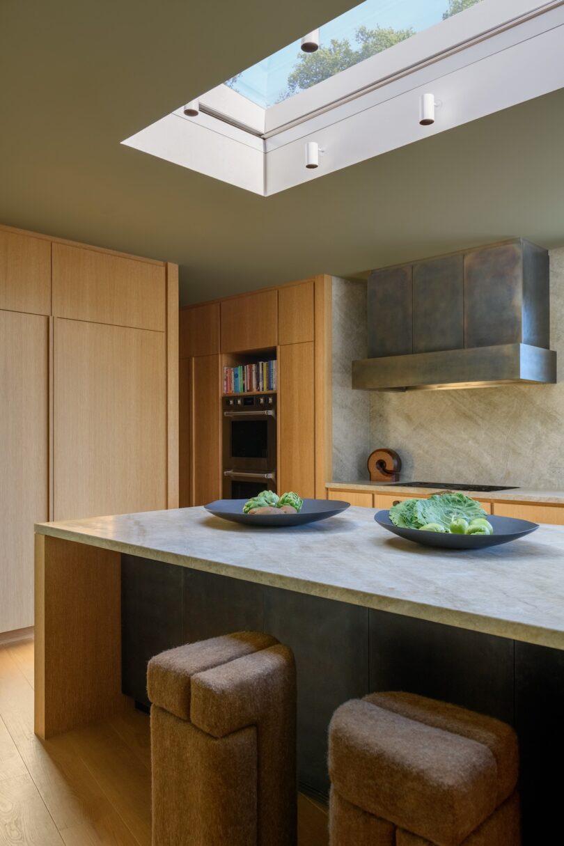 Modern kitchen with wood cabinets, marble countertops, two brown stools, and a skylight above. Plates with vegetables are on the island. Built-in appliances and shelves are visible.