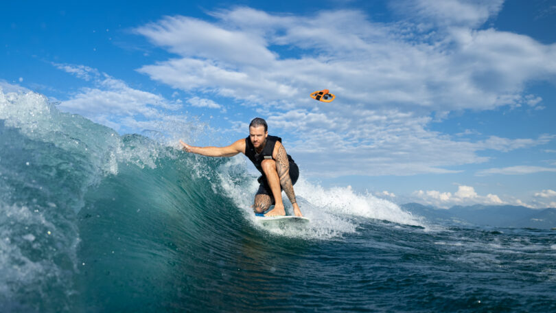 A man surf on the wave under the blue sky with scattered clouds; The small drone is visible to fly above the water in the background.
