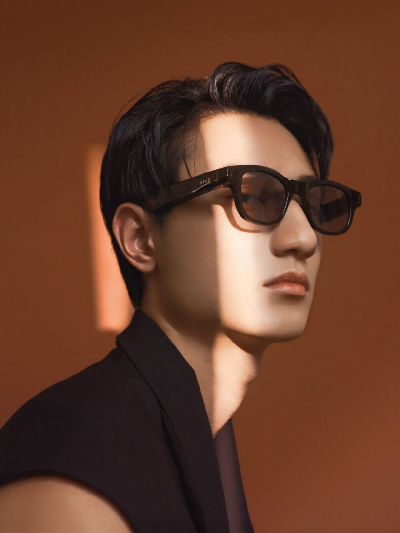 A person wearing black sunglasses and a dark outfit is lit by a narrow beam of sunlight against a brown background.