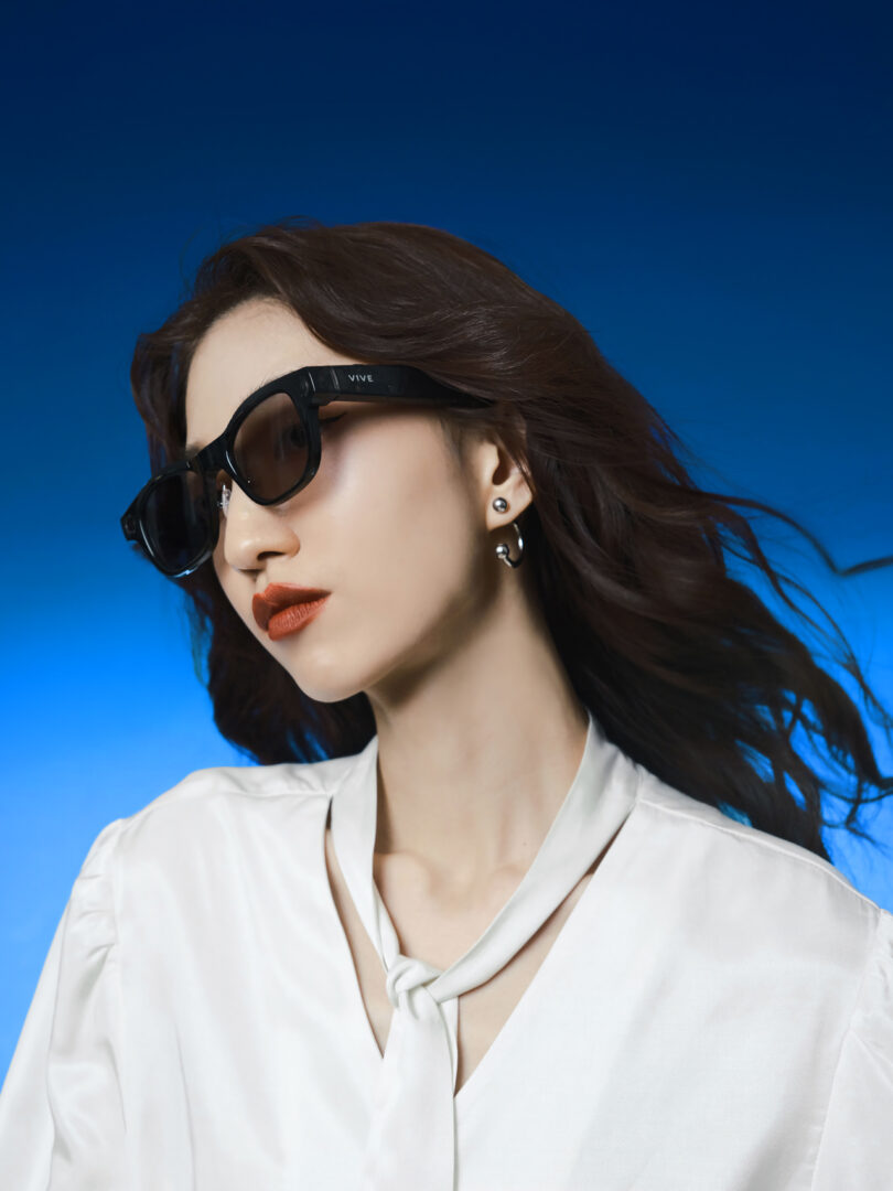 Woman with long dark hair wearing black sunglasses, white blouse, and earrings, posing against a gradient blue background.