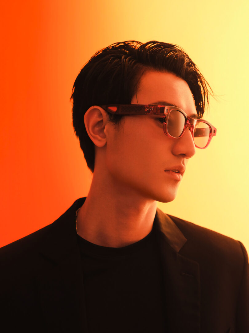 A young man wearing sunglasses and a black blazer looks to the side against a gradient orange and yellow background.