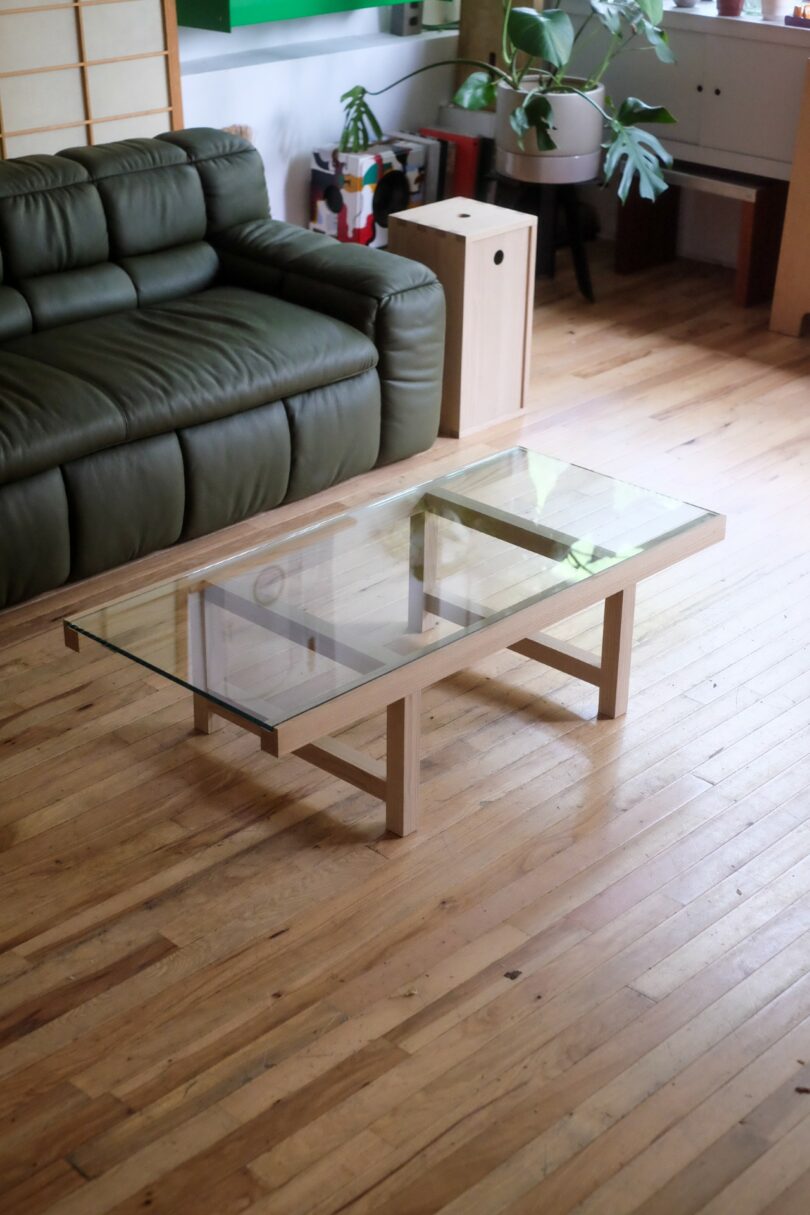 A glass coffee table with a light wood frame sits on a hardwood floor in a living room with a green sofa and several potted plants