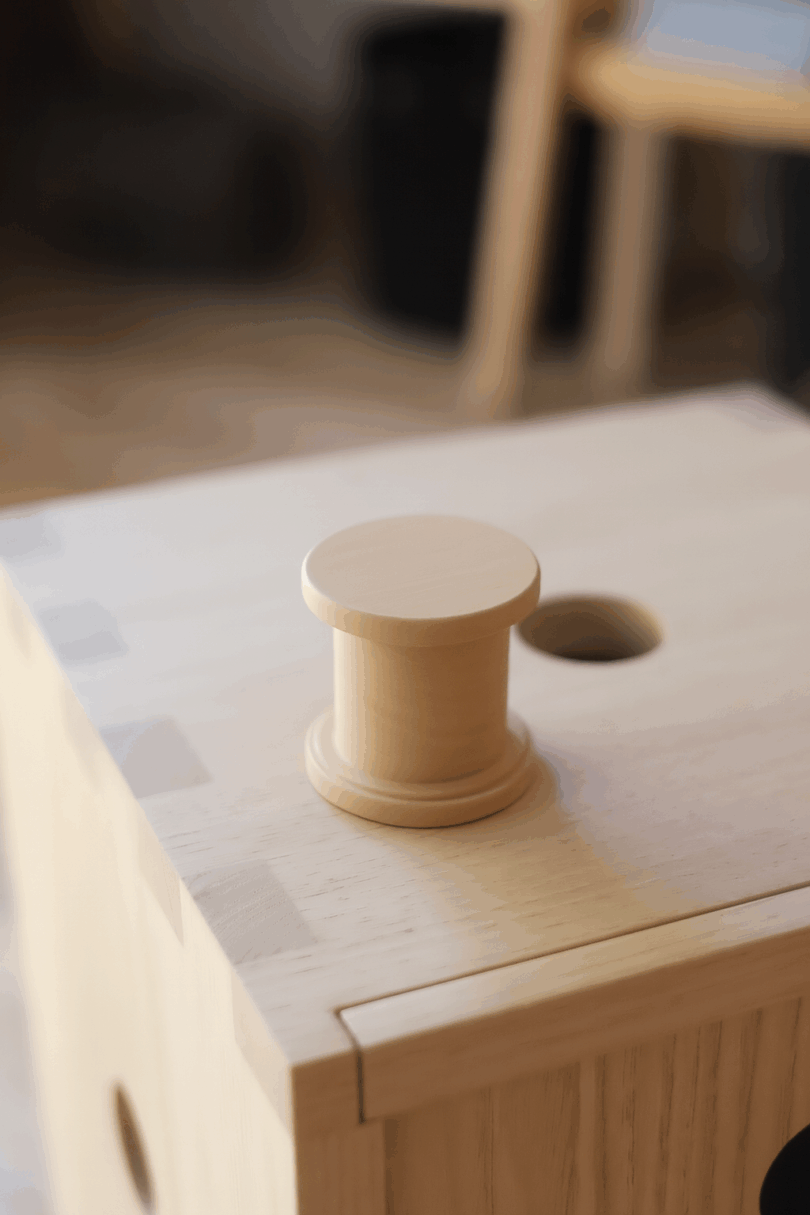A wooden spool-shaped object sits on top of a light wood box with a round hole, indoors, with blurred background furniture