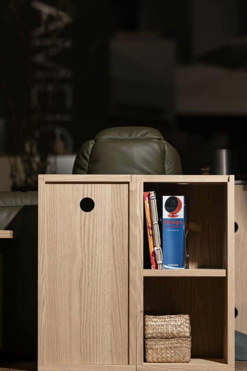 A light wood cabinet with a sliding door holds a rolled magazine, a book titled "Design," and two woven baskets on a lower shelf. A green chair is partially visible behind it
