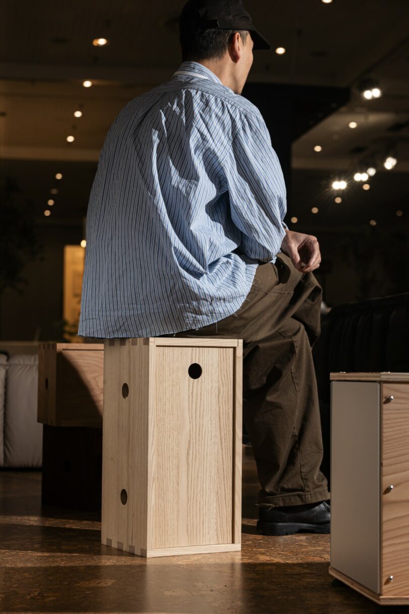 A person wearing a blue striped shirt and brown pants sits on a wooden stool in an indoor setting with warm lighting