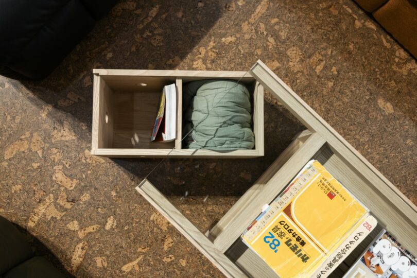 A wooden drawer containing a green blanket and a book is partially open beside a glass-topped table holding books and papers on a brown cork floor