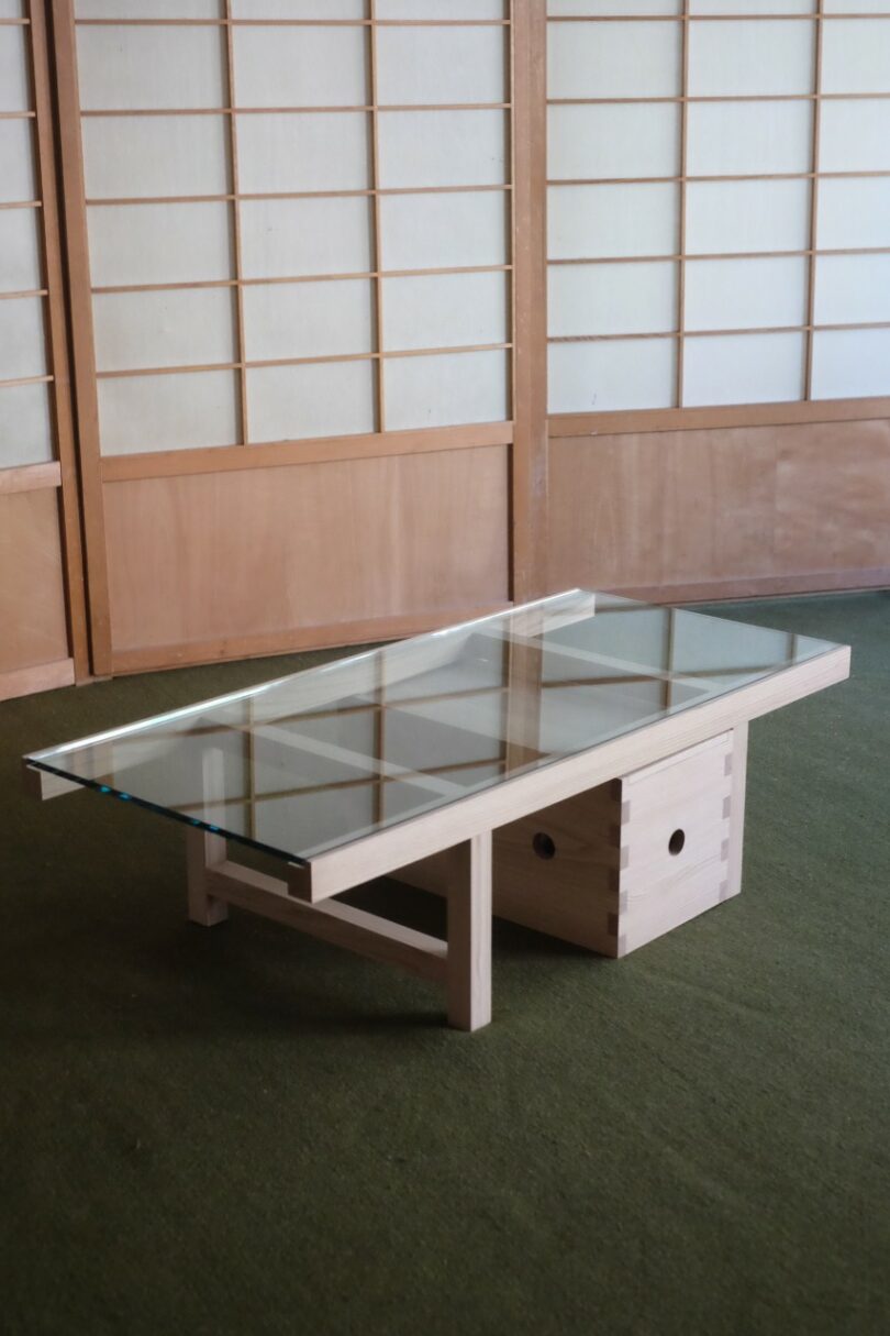 A rectangular glass-top coffee table with a light wood base sits on a green carpet in front of wooden shoji screen panels