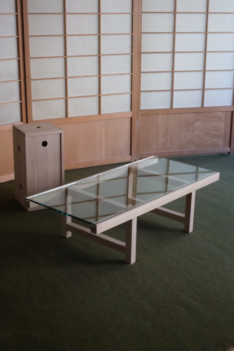 A glass-top coffee table with a wooden frame and a wooden box-shaped seat set on a green carpet in front of a shoji-style wall