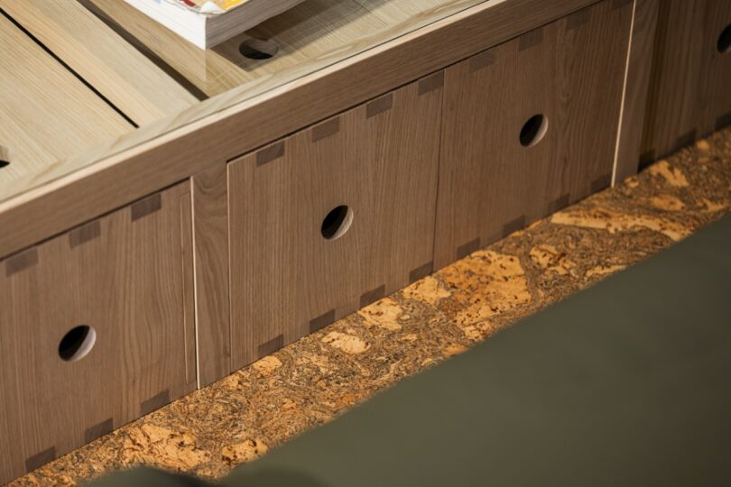 Close-up of wooden storage drawers with round cut-out handles, set against a cork floor and partially visible green upholstered furniture